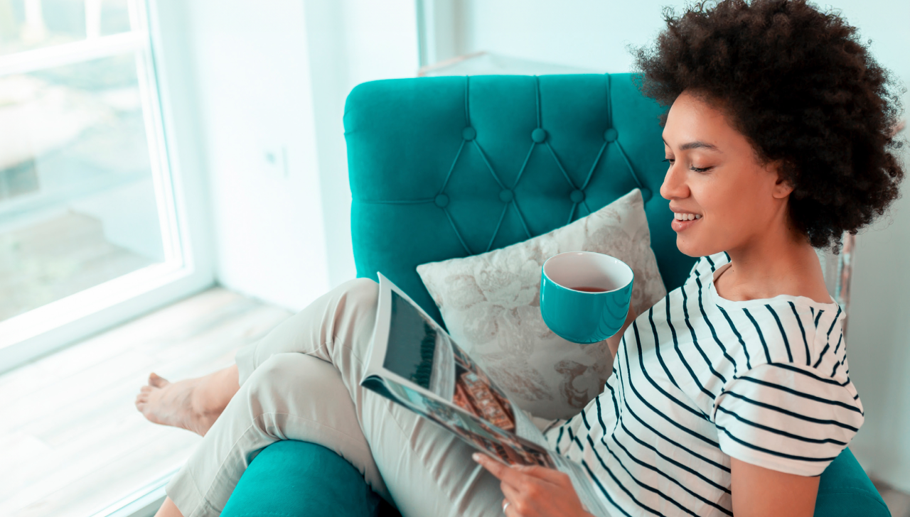 A woman sits in a chair reading a catalogue. She holds a teal mug of tea that matches the chair she is sitting in.