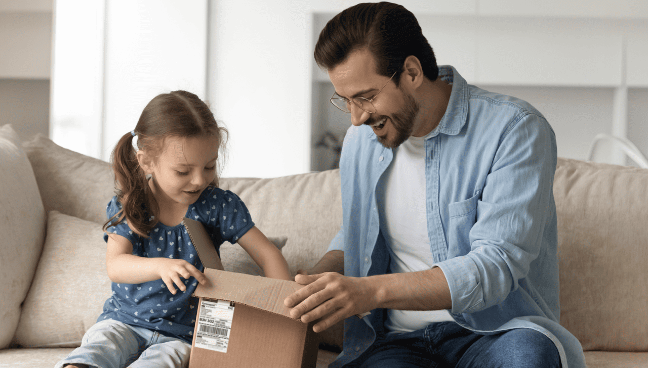 Adult and child sitting on a couch opening a small cardboard package together in a bright living room.