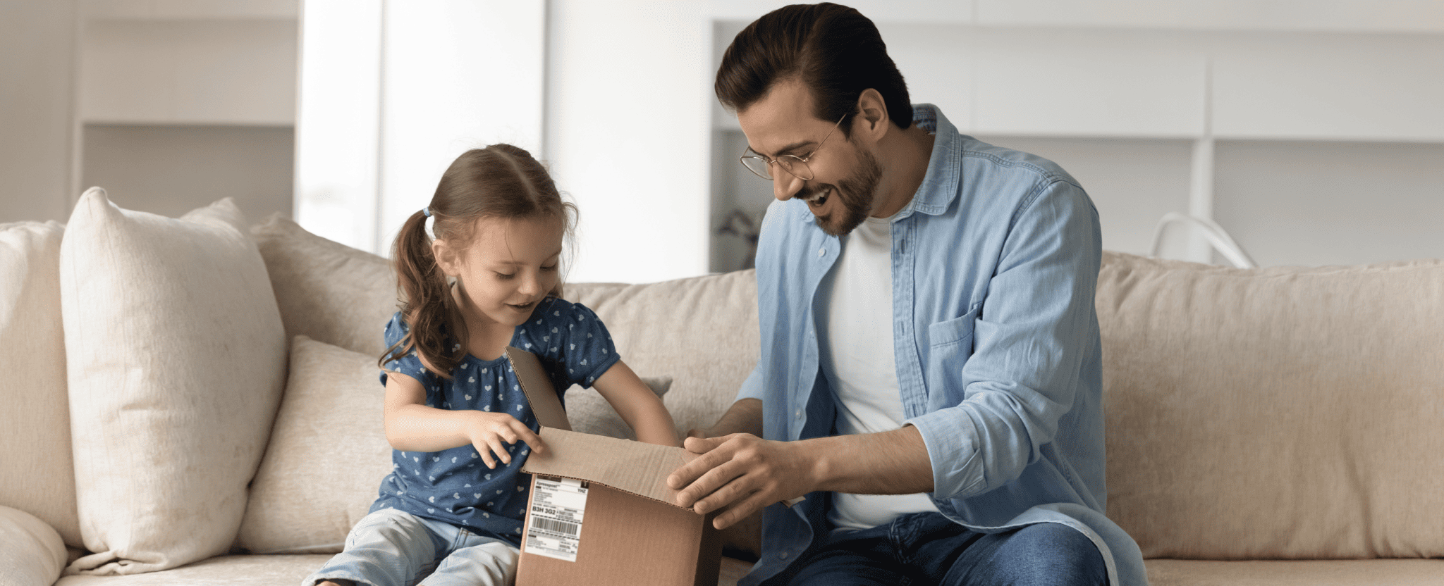 Adult and child sitting on a couch opening a small cardboard package together in a bright living room.