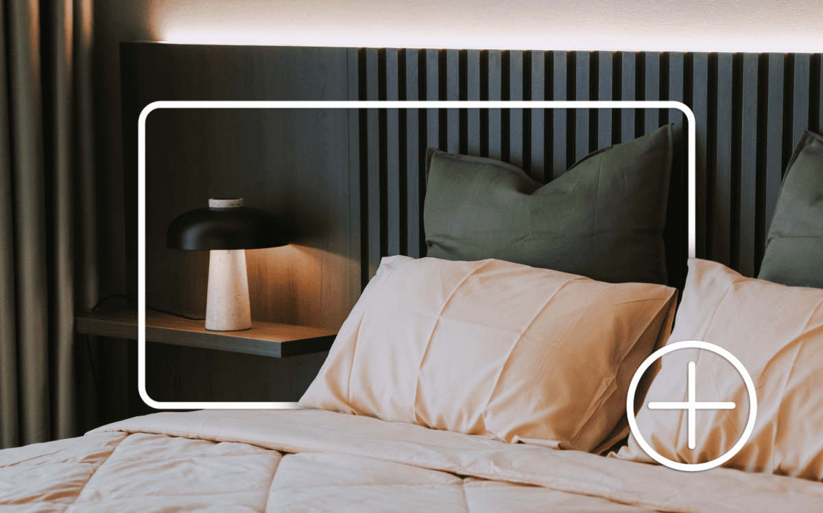 A bed with pillows, bedside table and lamp. Items are outlined by a white box and plus sign indicating adding to an online shopping cart.