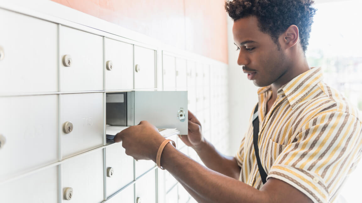 A man retrieves mail from a group mailbox compartment in an apartment building lobby.