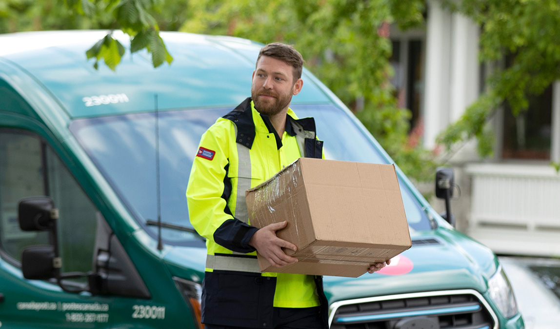 A Canada Post employee in a bright yellow jacket carries a package towards its destination. A hybrid Canada Post van is parked behind him.