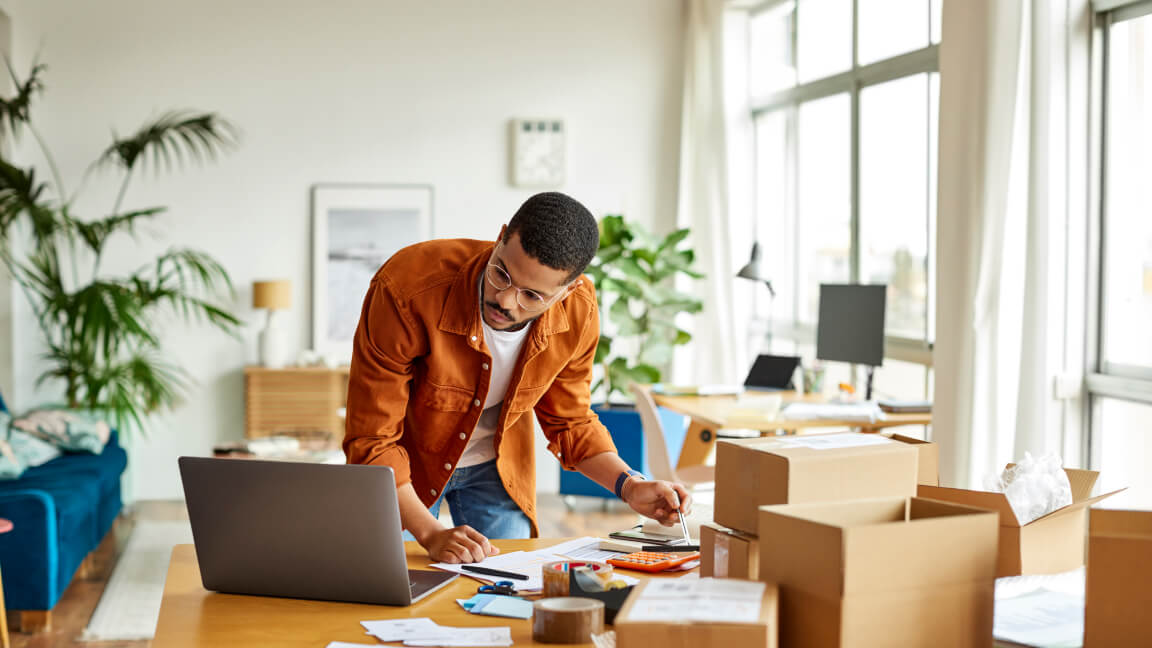 A man looks at a laptop while leaning over a table covered with small boxes and shipping supplies.