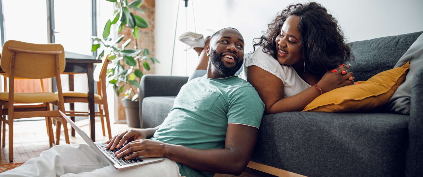 A man types on a laptop and leans against a couch a woman is sitting on. They smile at each other.