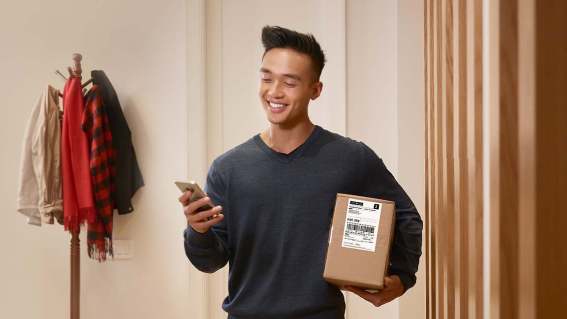 A young man smiles as he holds his cell phone in one hand and a parcel under his other arm.