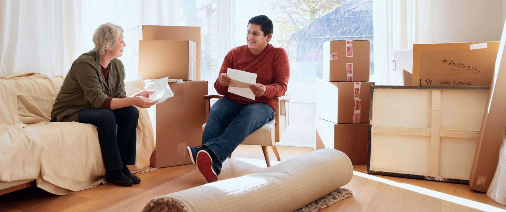 A woman sits holds papers and smiles at a young man who is sitting and holding papers. They are sitting together in a room full of moving boxes and items wrapped for moving.