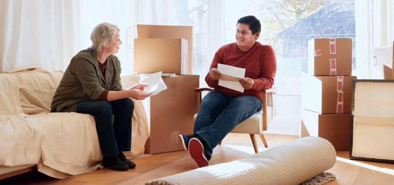 A woman sits holds papers and smiles at a young man who is sitting and holding papers. They are sitting together in a room full of moving boxes and items wrapped for moving.