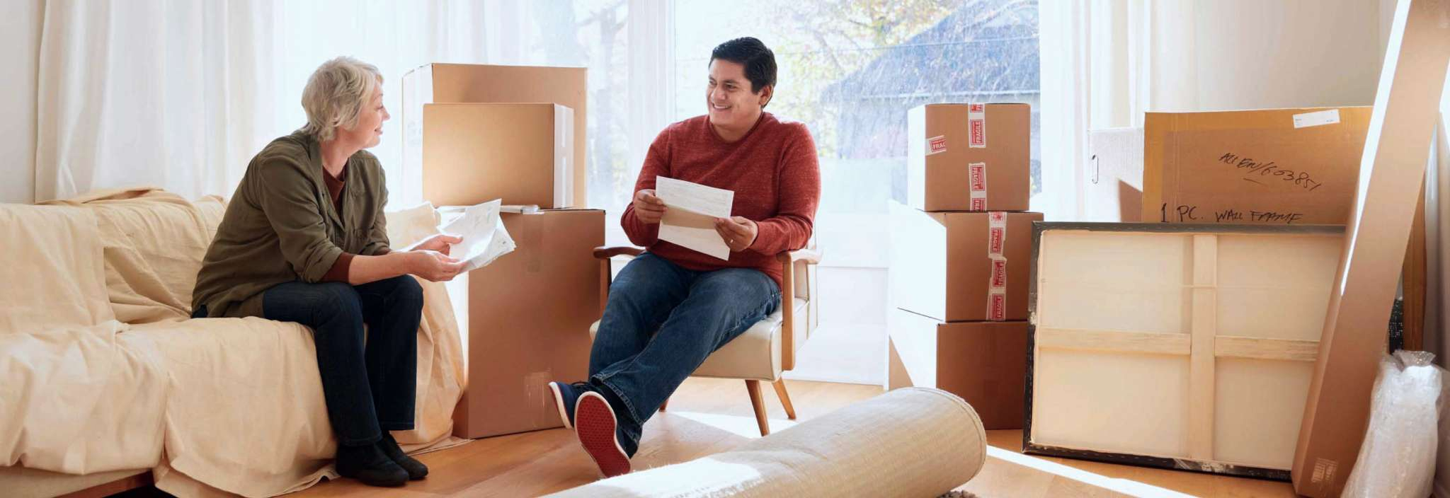 A woman sits holds papers and smiles at a young man who is sitting and holding papers. They are sitting together in a room full of moving boxes and items wrapped for moving.