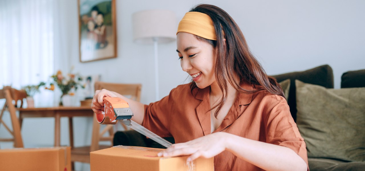 A young woman wearing a headband seals a shipping box with tape.